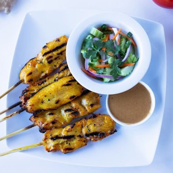 Grilled chicken satay skewers with peanut sauce and a small cucumber salad on a white plate.